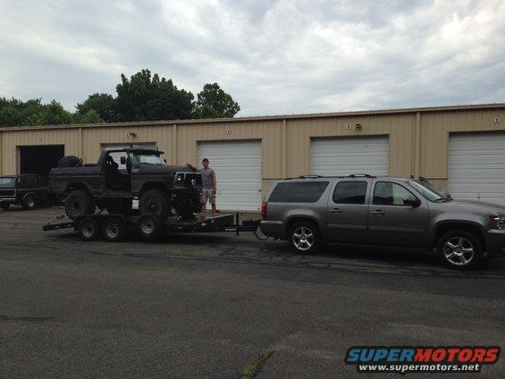 Black 1978 Ford Bronco on a triple-axle trailer, towed by a grey Chevrolet Suburban