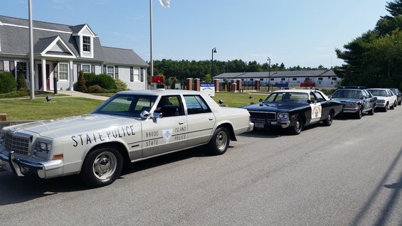 White police car with 'STATE POLICE RHODE ISLAND POLICE' text, parked in a line with other police cars