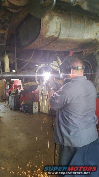 Man welding the rear axle of a rusty vehicle, sparks flying. A tire is visible