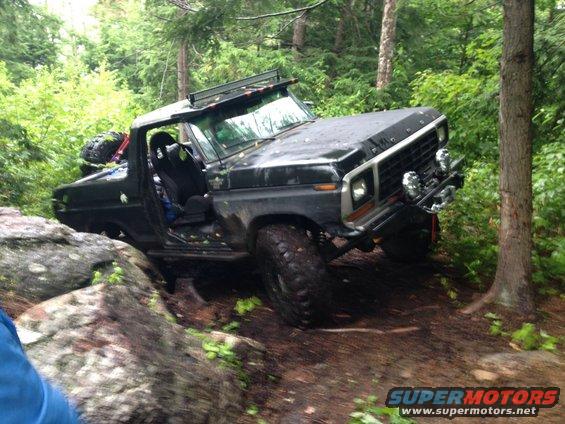 Black Ford Bronco on a rocky, wooded trail. It has a light bar on top and a spare tire mounted on the back
