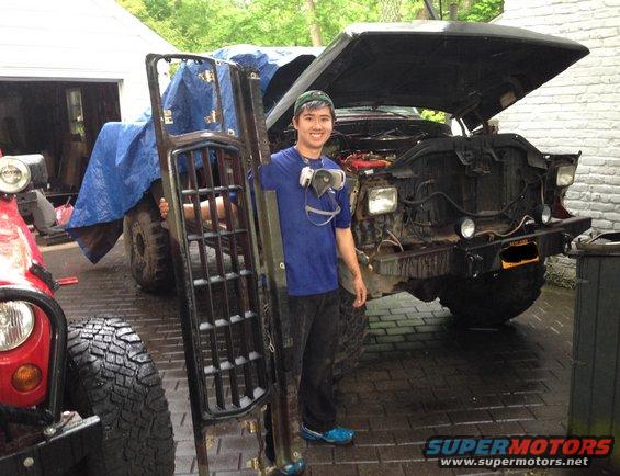 Man holding a large grille in front of a dark-colored truck with its hood open. A red Jeep is partially visible on the left