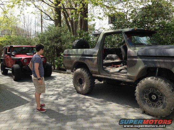 Camouflaged Ford Bronco with doors removed, parked next to a red Jeep Wrangler. A person stands between them