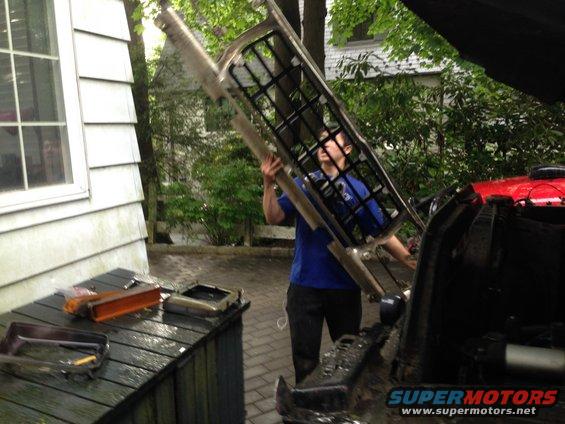 Man holding a large metal grille part over an open vehicle hood