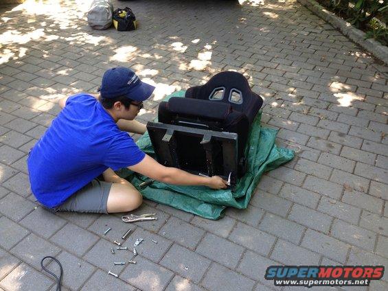 Guy kneeling on paver driveway, working on a racing seat on a green tarp. Bolts and tools are on the ground