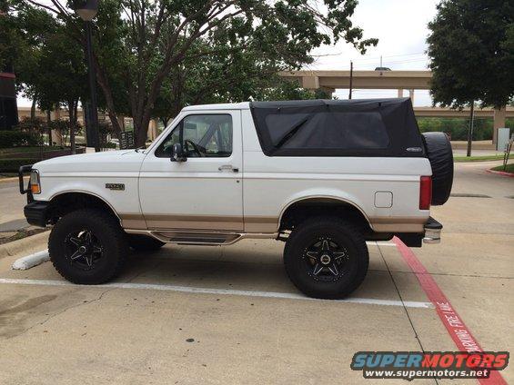 White Ford Bronco with a black soft top and spare tire on the back, parked on pavement