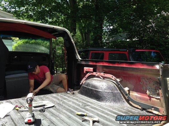 Guy fixing up the bed of an old red pickup truck, another red vehicle is parked behind it