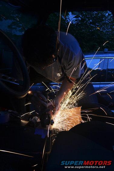 Interior of a dark vehicle with a person grinding metal, sparks flying. A blue car is visible through the window