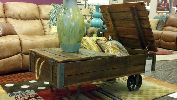 Wooden storage chest with rope handles and metal accents, open lid revealing pillows, on wheels, next to a leather sofa
