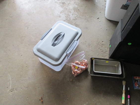 Plastic storage bin with lid, bag of copper fittings, and metal baking pans on concrete floor