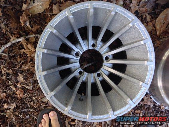 Close-up of a silver alloy car wheel with 15 spokes, resting on dry leaves. A person's foot in a sandal is visible at the bottom left