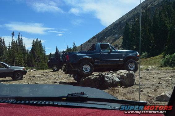 Dark blue pickup truck with off-road tires on rocks, other vehicles and trees in background