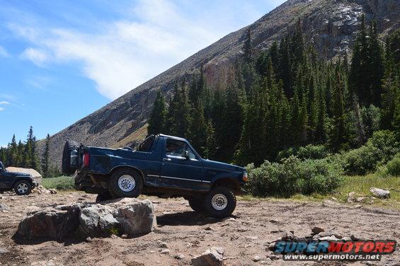 Dark blue Ford Bronco on rocky terrain with a mountain and trees in background. Another vehicle is partially visible to the left