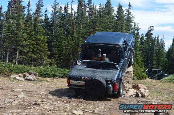 Dark blue Ford Bronco is stuck on a rock with its rear tire on the ground. Another Jeep is visible in the background