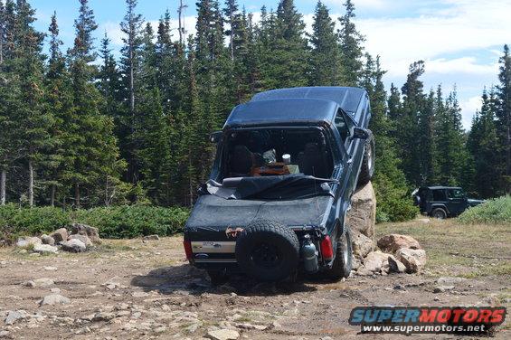 Dark blue SUV with a spare tire on the back, tilted on a rock, with another dark SUV in the background