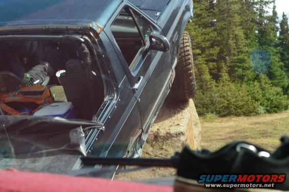 1983 Ford Bronco with driver's door open, climbing over a large rock off-road