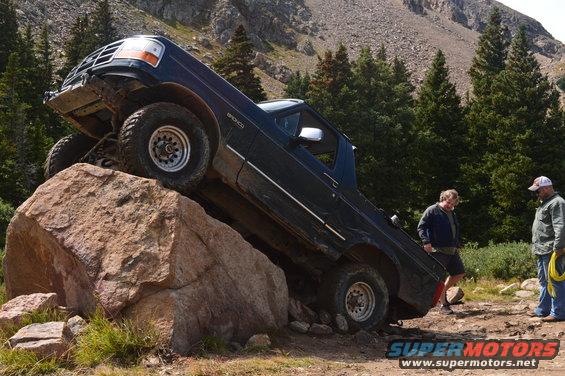 Dark blue Ford Bronco climbing a large rock on a trail, two men watching