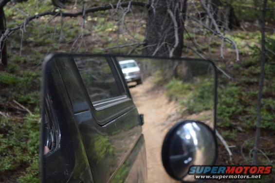 White SUV seen in side mirror on a dirt road, surrounded by trees