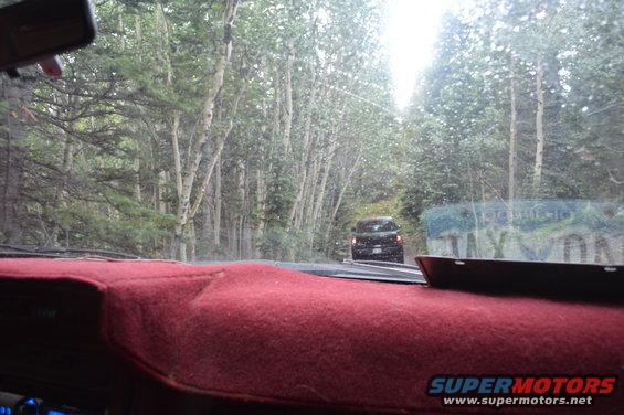 Interior view from a car driving on a dirt road through a forest, with another dark SUV ahead