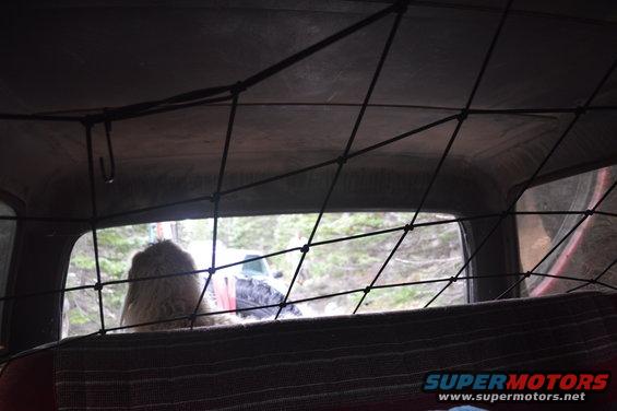 Interior view from a vehicle looking out the rear window at a wooded area and another vehicle