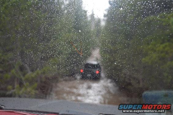 View through a wet windshield of a dark SUV driving on a muddy, water-filled trail between trees