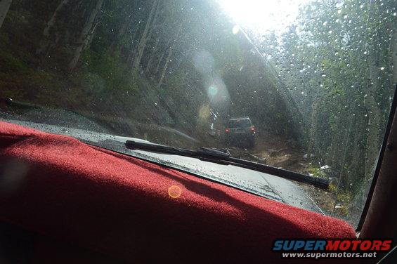 View from inside a vehicle looking out at another vehicle on a muddy, wooded trail