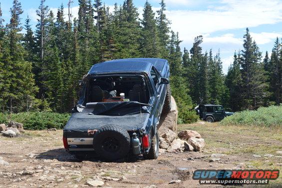 1983 Ford Bronco climbing over a rock on a dirt trail with trees and another Jeep in the background