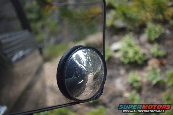 Side mirror reflection shows a dirt road and trees. The vehicle's side panel is visible