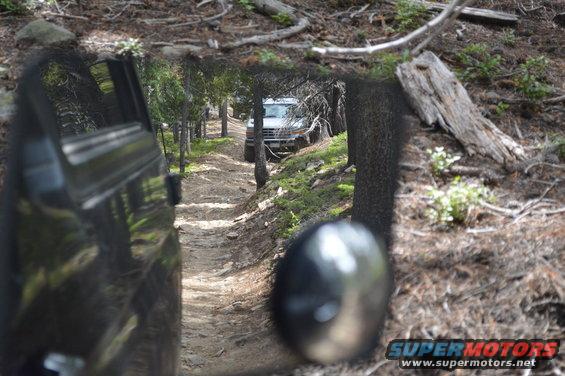 White SUV on dirt road, seen through reflection of another vehicle