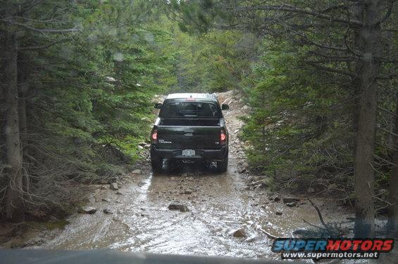 Black pickup truck driving on a muddy, rocky forest road with trees on both sides