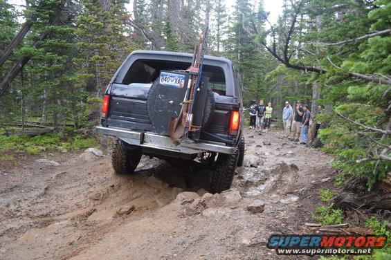Black 1983 Ford Bronco driving on a rocky, muddy trail with a shovel and jack mounted on the back. People watch from the side of the trail
