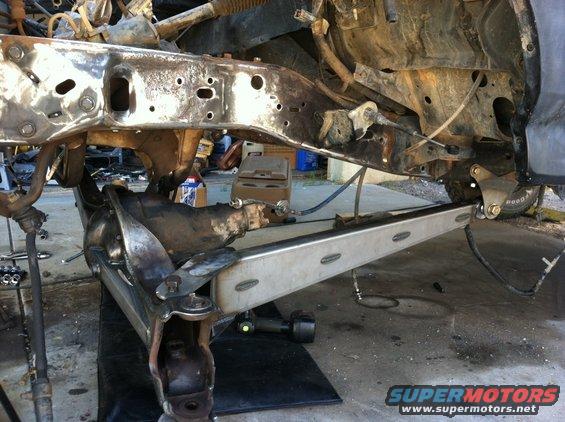Vehicle frame and suspension parts, possibly from a truck or SUV, in a workshop. Some parts are rusty, others appear recently worked on