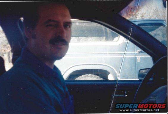 Man with mustache winks while sitting in driver's seat. Another vehicle is visible through the window