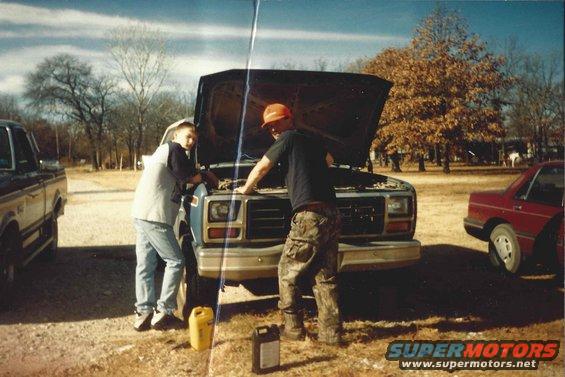 Two guys looking at the engine of a blue truck with the hood up. Another truck and a red car are nearby
