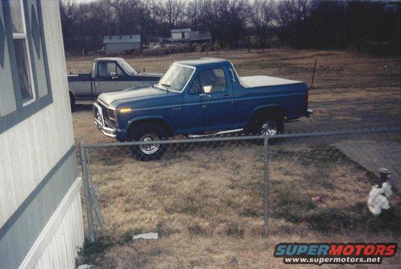 Blue 1980s Ford Bronco with a white soft top, parked in a grassy yard next to a trailer home