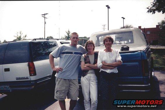 Three people stand between a white Chevrolet SUV and a blue pickup truck with a soft top