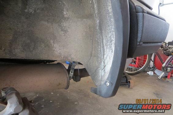 Car undercarriage showing exhaust pipe and wheel well liner. A bicycle is visible in the background