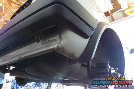Underside view of a car's rear bumper and wheel well, showing rust and suspension components