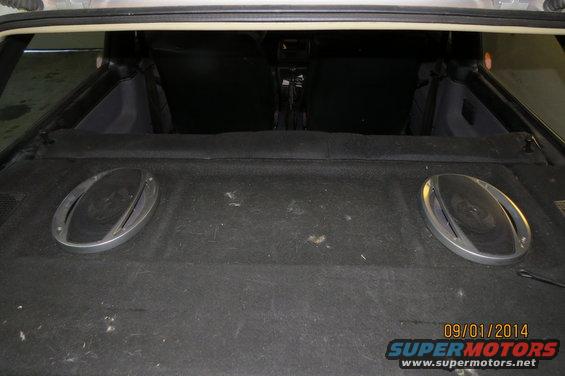 Rear cargo area of a car showing two oval speakers mounted on the carpeted floor. Interior seats are visible in the background