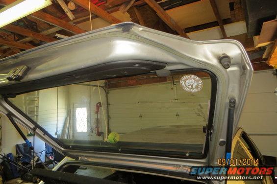 Interior of a silver vehicle's rear hatch, showing the glass and surrounding trim. A sticker is visible on the glass