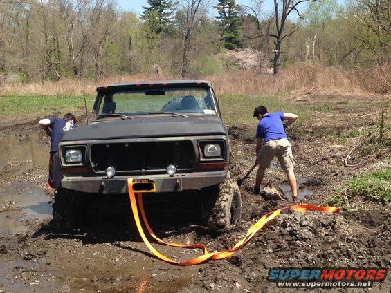 Ford Bronco stuck in mud, two people trying to free it with a strap and shovel
