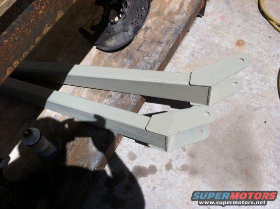 Two painted metal suspension components on a workbench, with a brake rotor and can of spray paint nearby