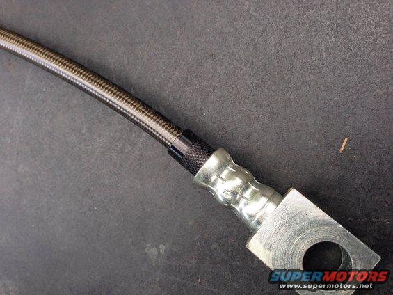 Close-up of a braided brake hose with metal fittings. The hose has a carbon fiber pattern, and the fittings are silver metal