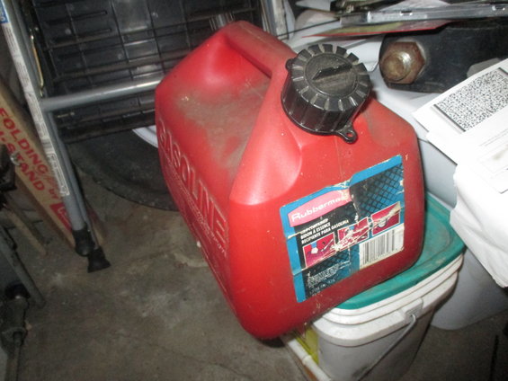 Red Rubbermaid gas can with black cap, sitting on buckets. Some boxes and a folding stand are in the background