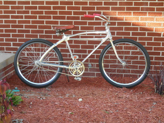 Chrome frame bicycle with red grips and brown leather seat, parked against a brick wall