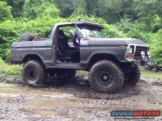 Dark gray Ford Bronco with off-road tires driving through mud, doors removed, spare tire on back