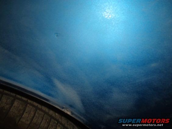 Close-up of a dent on a blue car's body panel, with a tire visible below