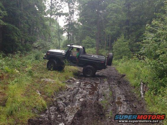Black Ford Bronco with spare tire on rear, driving on a muddy forest trail. The Bronco appears to be a 1978 model