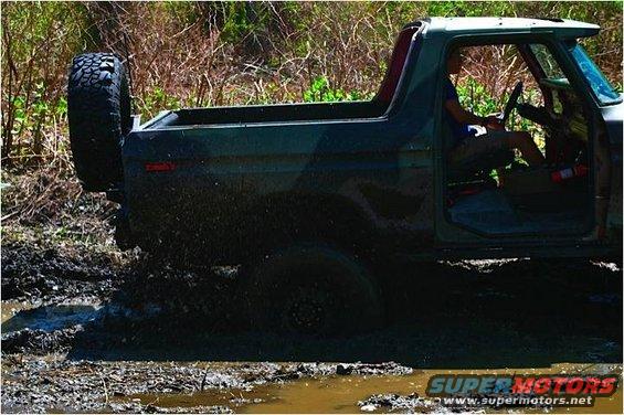 Dark green 1978 Ford Bronco driving through mud, spare tire on rear, driver visible