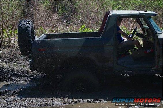 Dark green 1979 Ford Bronco side view, driving through mud with spare tire mounted on rear