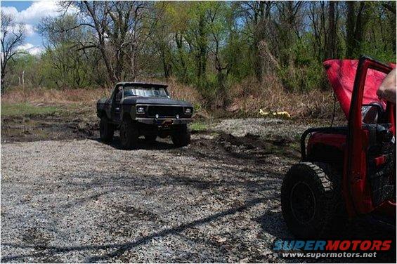 Black pickup truck with lifted suspension in a muddy area; red jeep in foreground